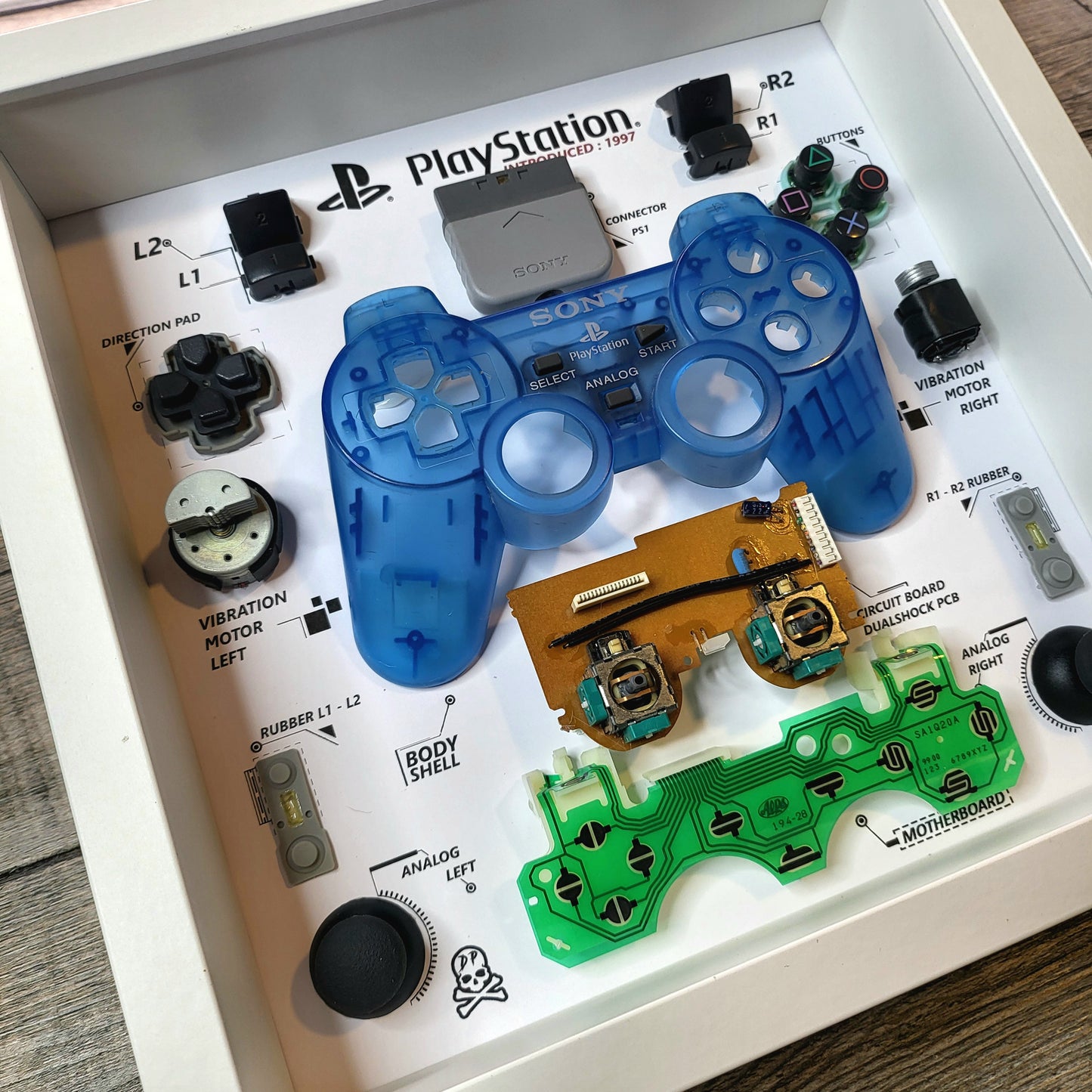 Components of a disassembled controller of PlayStation Ps1 Dualshock in a blue limited edition inside a white frame 27x27 cm with a protective polycarbonate transparent cover on the front