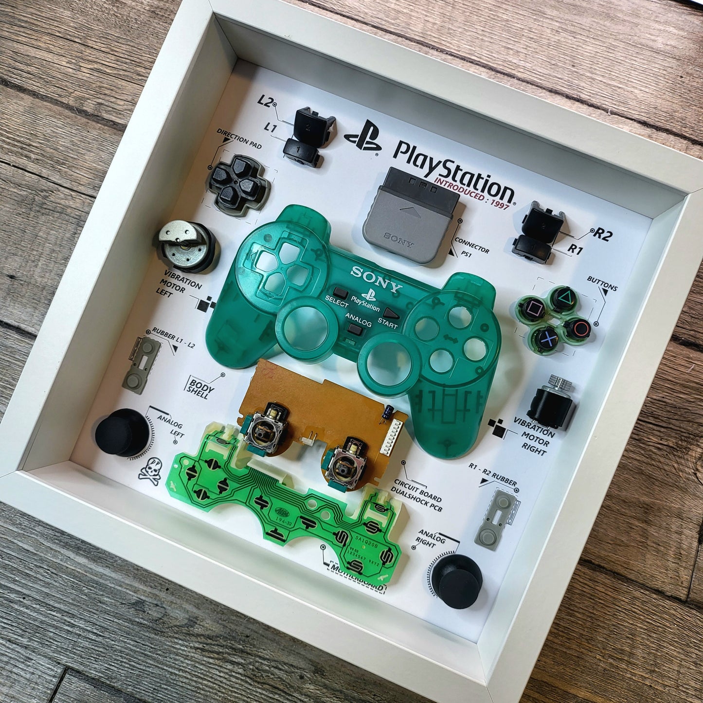 Components of a disassembled controller of PlayStation Ps1 Dualshock in a green limited edition inside a white frame 27x27 cm with a protective polycarbonate transparent cover on the front