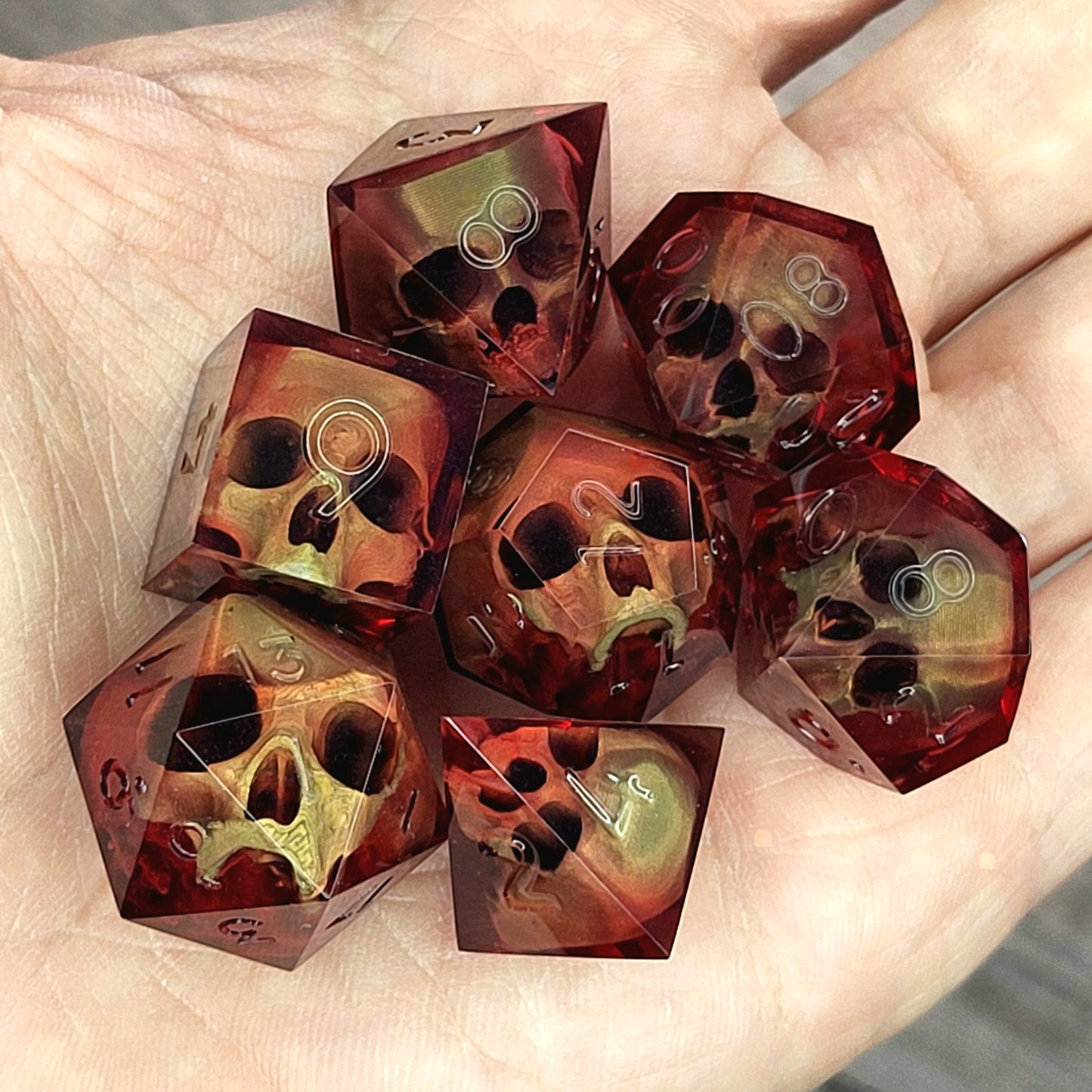 Handmade translucent red resin dice set with embedded skull designs resting in a hand. Gothic RPG polyhedral dice for Dungeons & Dragons and tabletop gaming with dark fantasy aesthetic