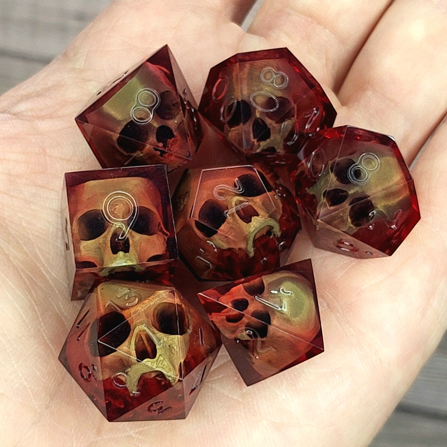 Handmade translucent red resin dice set with embedded skull designs resting in a hand. Gothic RPG polyhedral dice for Dungeons & Dragons and tabletop gaming with dark fantasy aesthetic