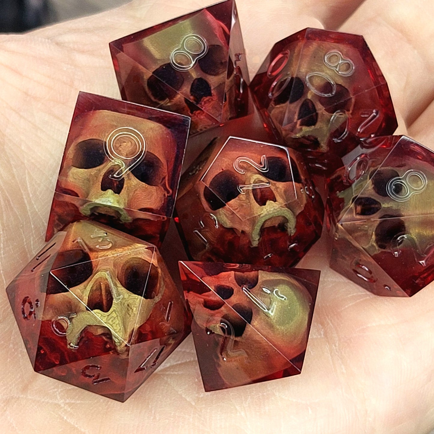 Handmade translucent red resin dice set with embedded skull designs resting in a hand. Gothic RPG polyhedral dice for Dungeons & Dragons and tabletop gaming with dark fantasy aesthetic