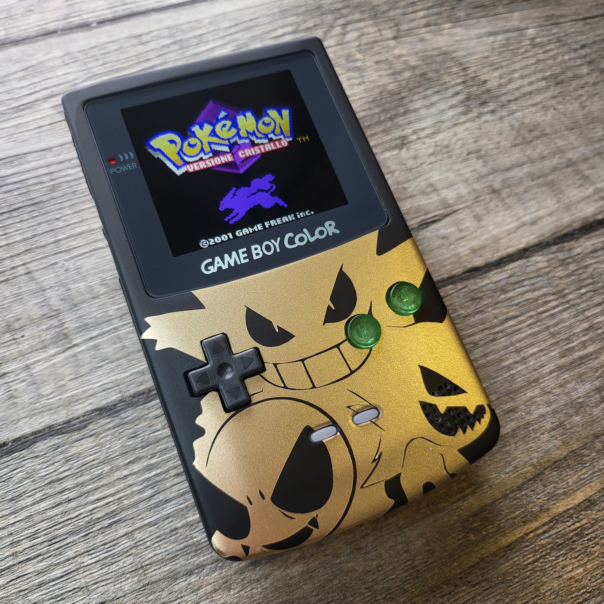 A customized Nintendo Gameboy color with black and gold Gengar pokemon on the front cover and backlit display