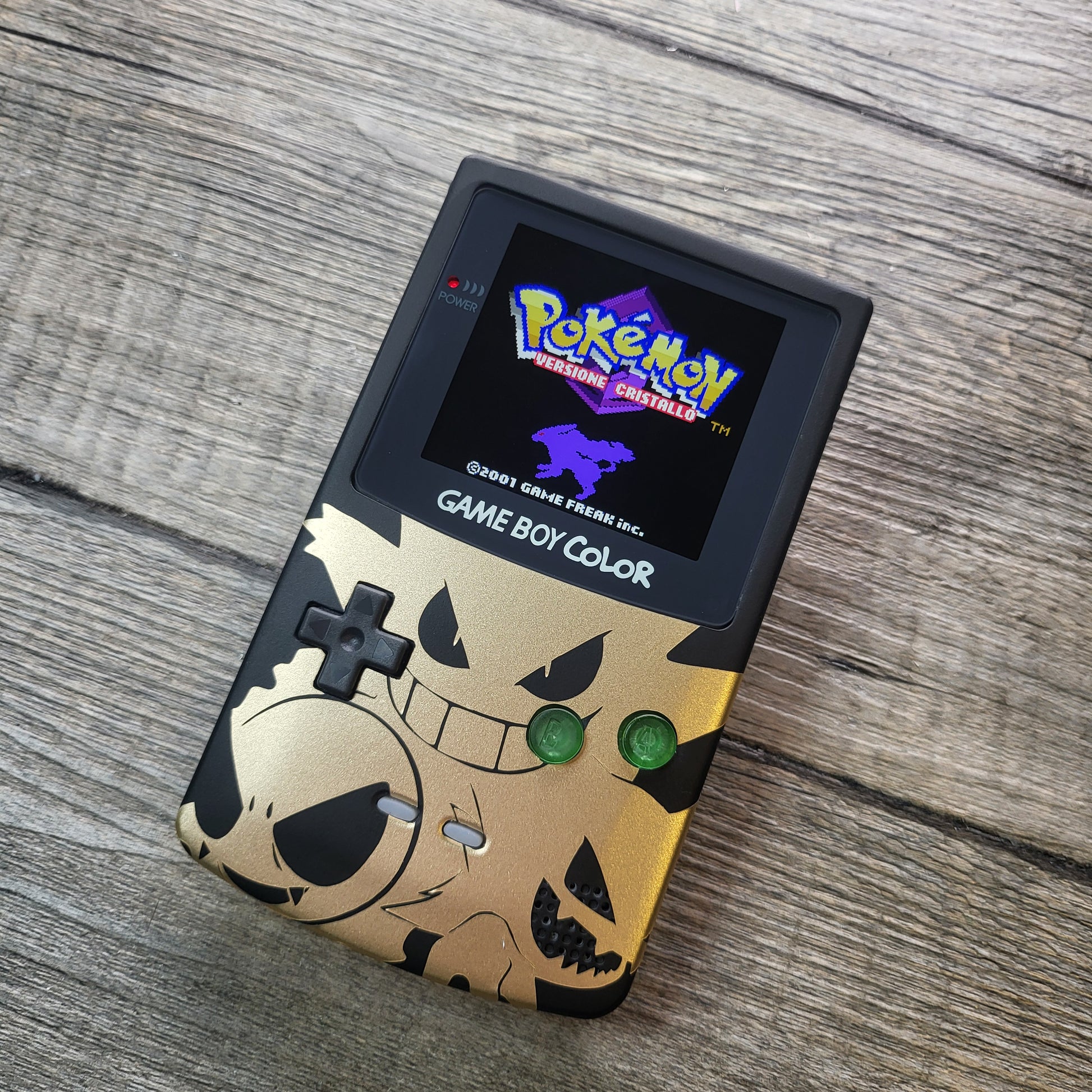 A customized Nintendo Gameboy color with black and gold Gengar pokemon on the front cover and backlit display
