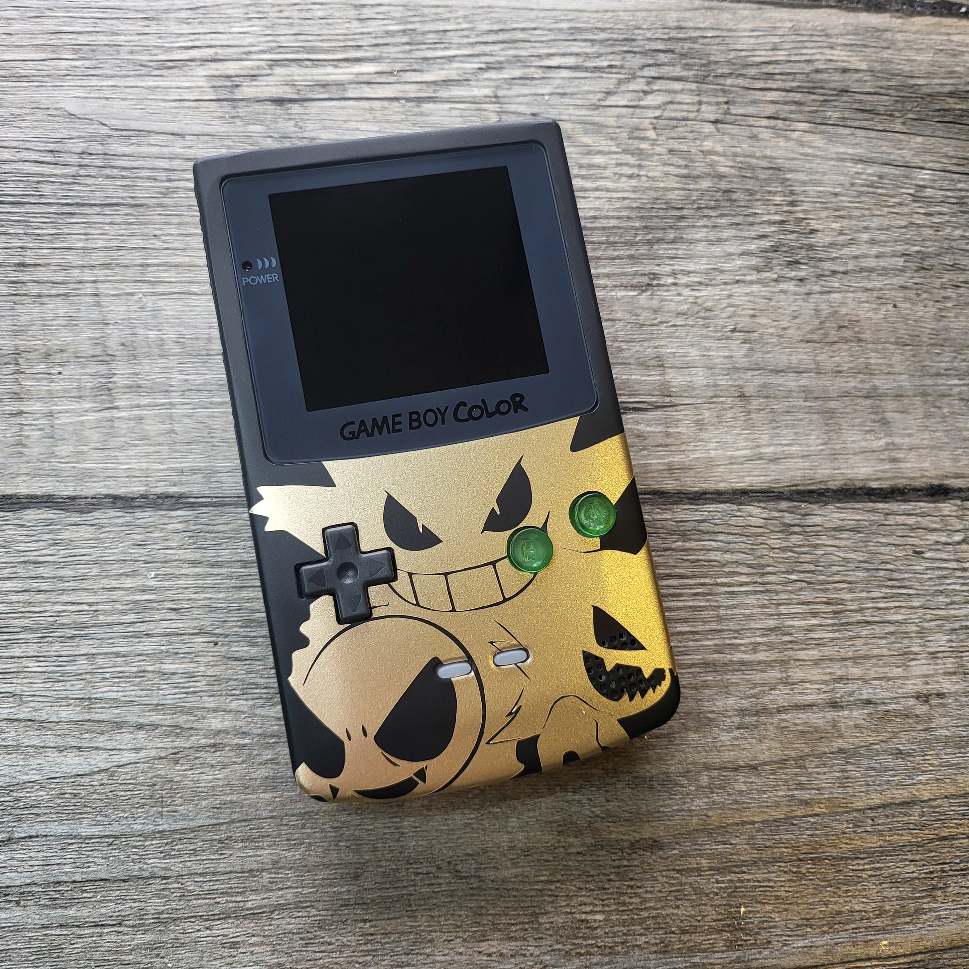 A customized Nintendo Gameboy color with black and gold Gengar pokemon on the front cover and backlit display