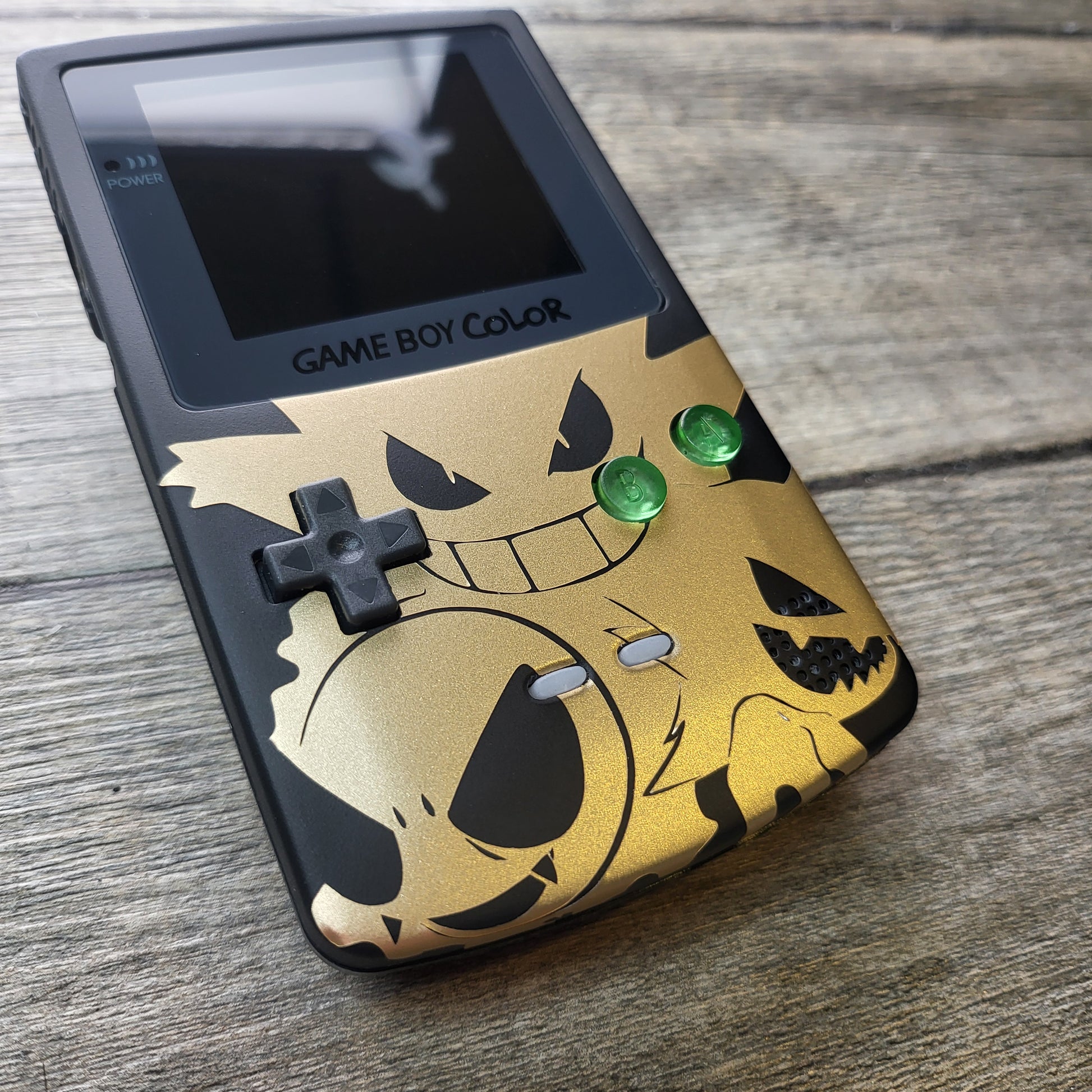 A customized Nintendo Gameboy color with black and gold Gengar pokemon on the front cover and backlit display