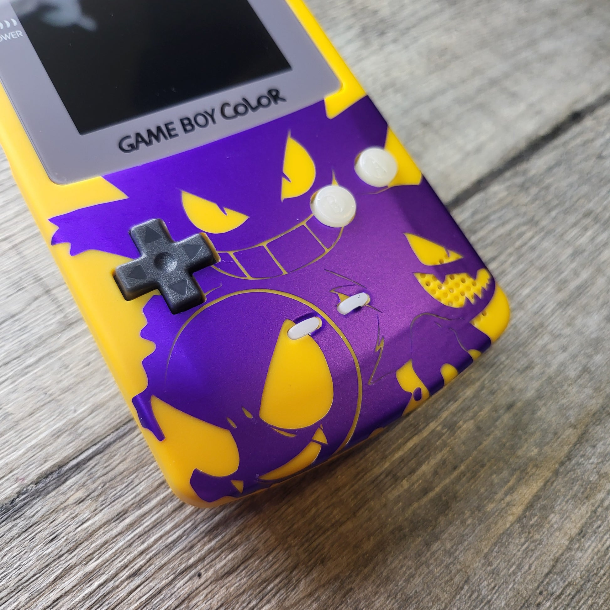 A customized Nintendo Gameboy color with yellow and purple Gengar pokemon on the front cover and backlit display