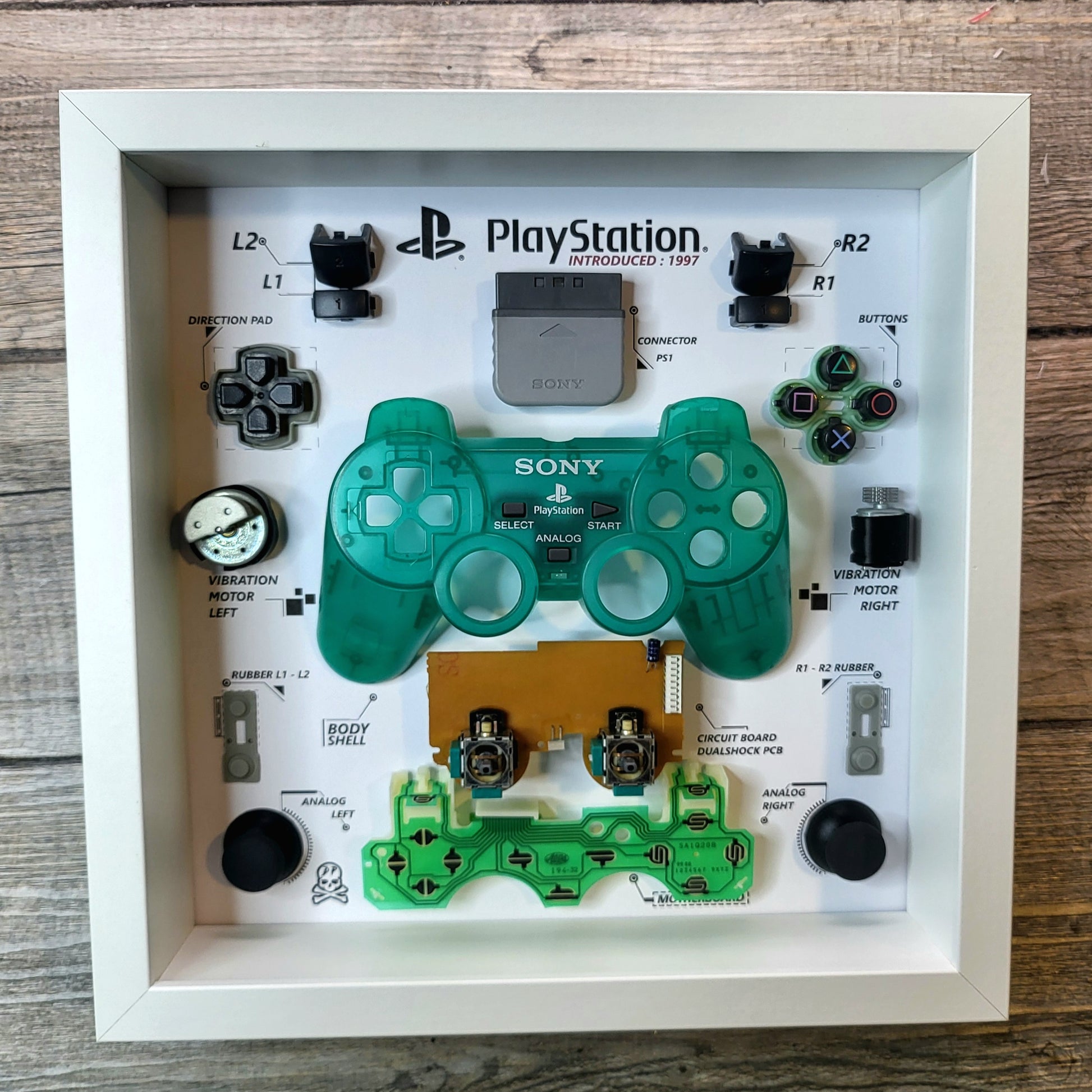 Components of a disassembled controller of PlayStation Ps1 Dualshock inside a white frame 27x27 cm with a protective polycarbonate transparent cover on the front in a green limited edition