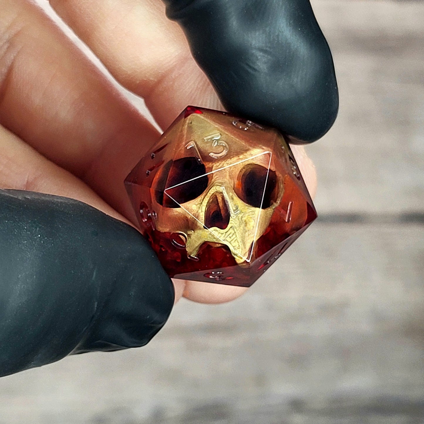 Handmade translucent red resin D20 dice with embedded skull design, held between black-gloved fingers. Gothic tabletop gaming dice with dark fantasy aesthetic for Dungeons & Dragons and RPG players