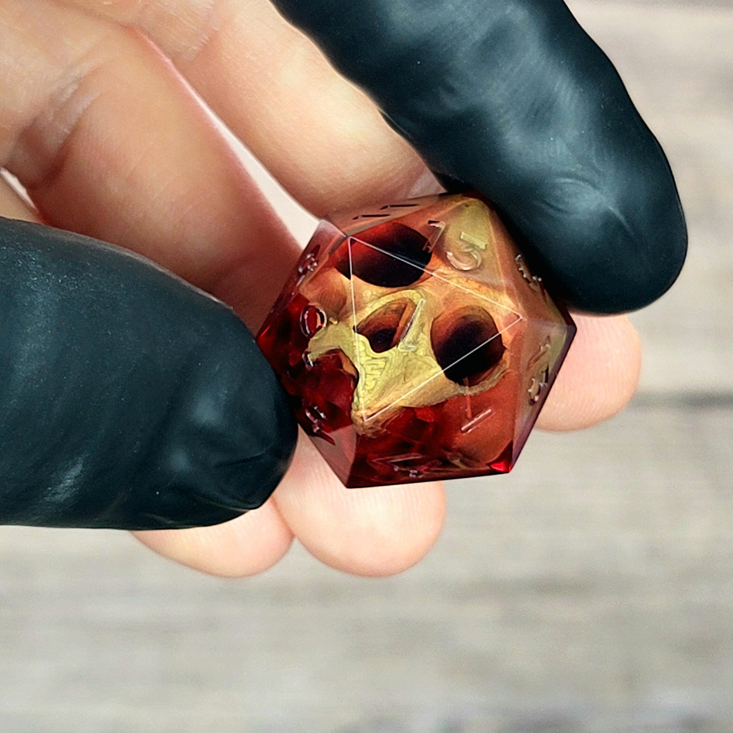 Handmade translucent red resin D20 dice with embedded skull design, held between black-gloved fingers. Gothic tabletop gaming dice with dark fantasy aesthetic for Dungeons & Dragons and RPG players