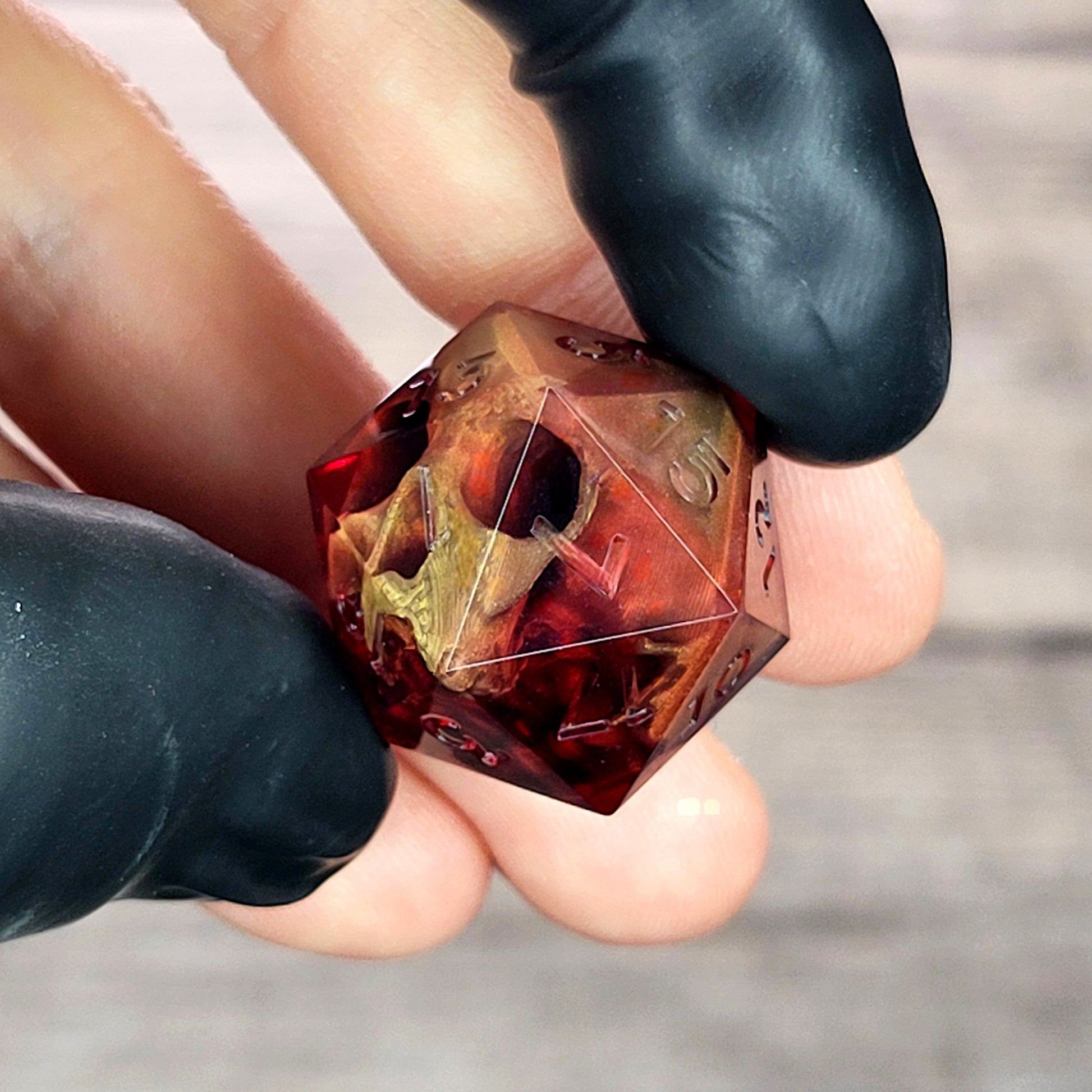 Handmade translucent red resin D20 dice with embedded skull design, held between black-gloved fingers. Gothic tabletop gaming dice with dark fantasy aesthetic for Dungeons & Dragons and RPG players