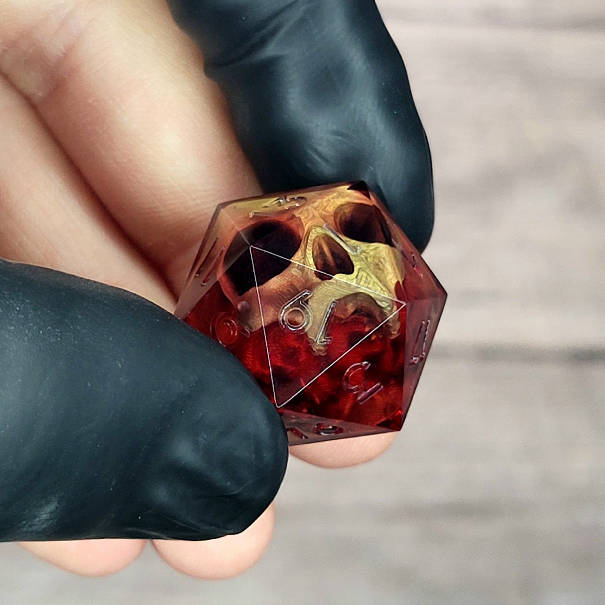 Handmade translucent red resin D20 dice with embedded skull design, held between black-gloved fingers. Gothic tabletop gaming dice with dark fantasy aesthetic for Dungeons & Dragons and RPG players