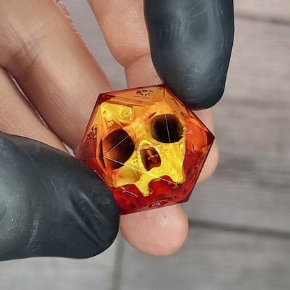 Red translucent resin D20 dice with a gold skull embedded inside, held between black-gloved fingers. Handmade gothic RPG dice for Dungeons & Dragons and dark fantasy tabletop gaming