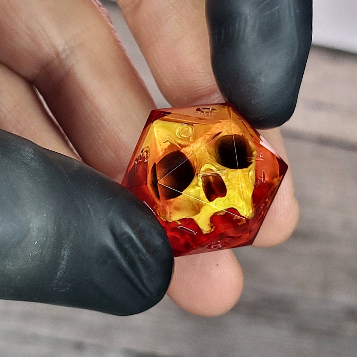 Red translucent resin D20 dice with a gold skull embedded inside, held between black-gloved fingers. Handmade gothic RPG dice for Dungeons & Dragons and dark fantasy tabletop gaming