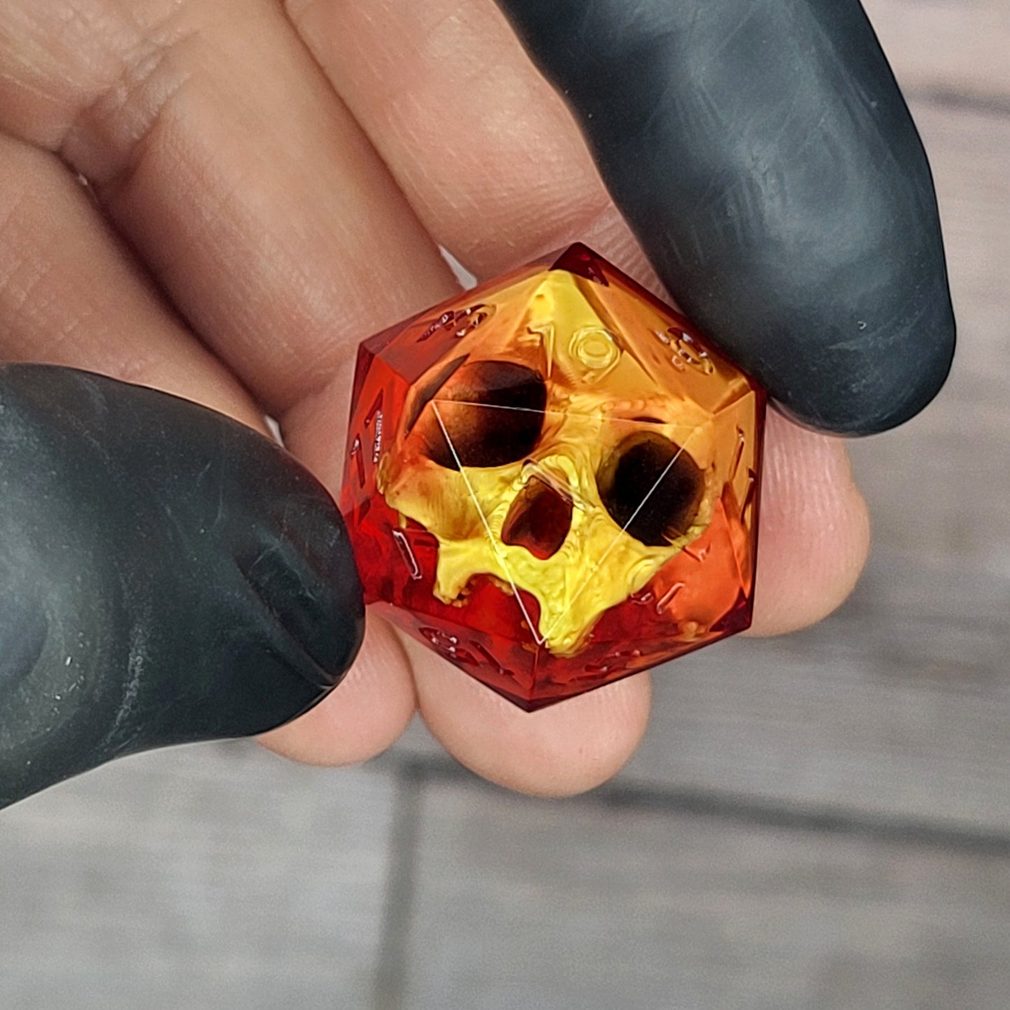 Red translucent resin D20 dice with a gold skull embedded inside, held between black-gloved fingers. Handmade gothic RPG dice for Dungeons & Dragons and dark fantasy tabletop gaming