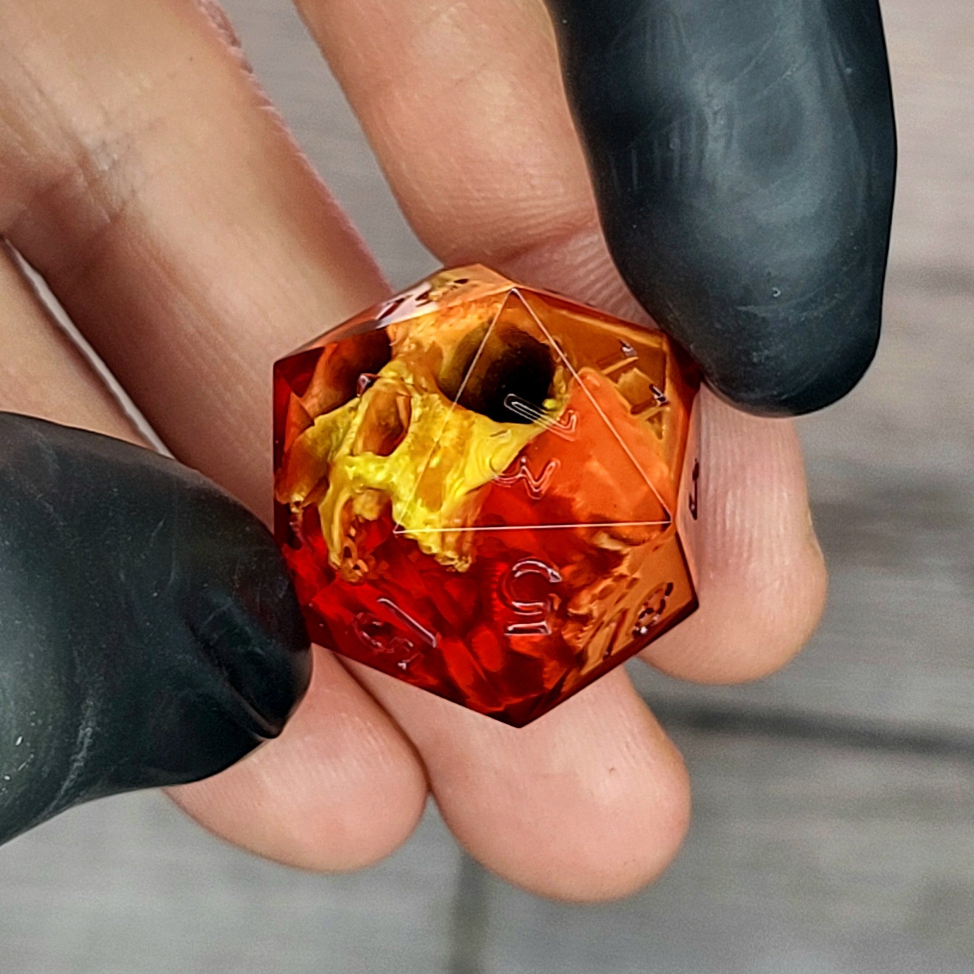 Red translucent resin D20 dice with a gold skull embedded inside, held between black-gloved fingers. Handmade gothic RPG dice for Dungeons & Dragons and dark fantasy tabletop gaming