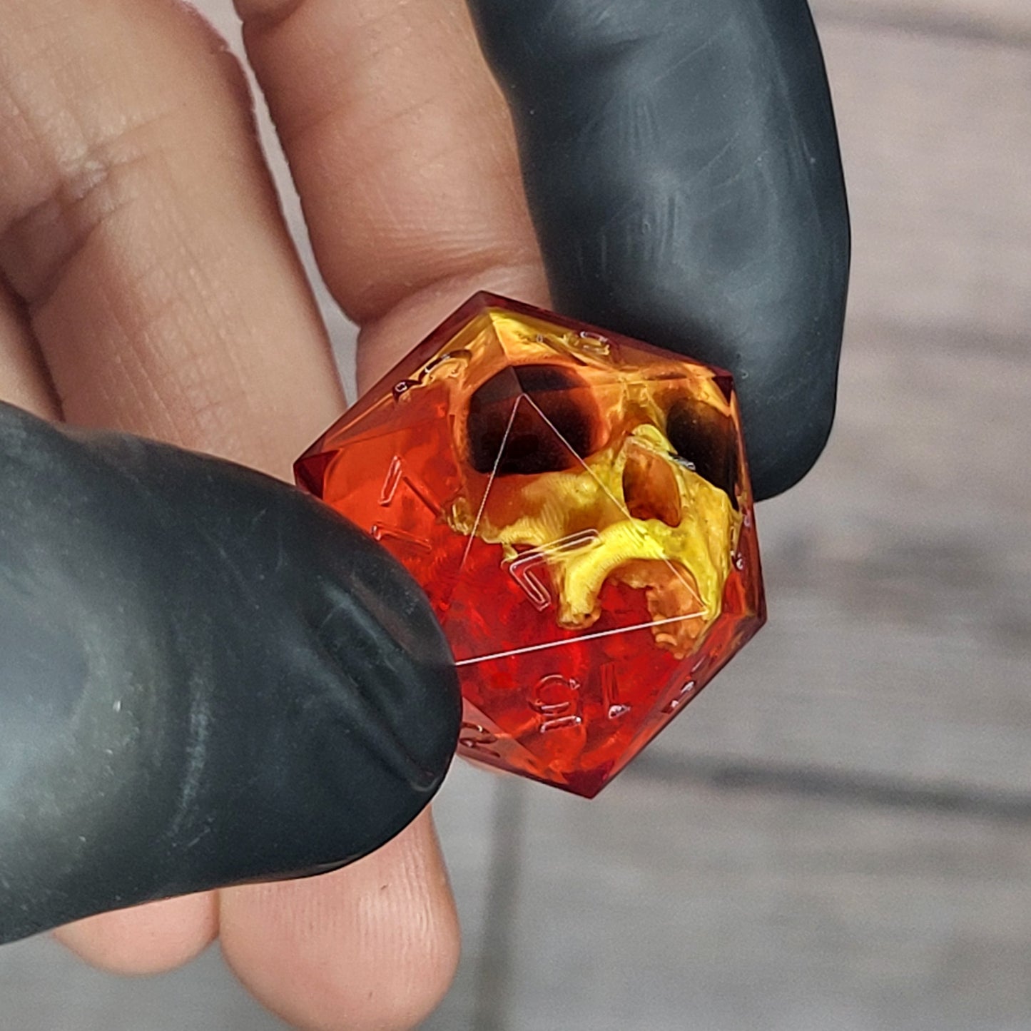 Red translucent resin D20 dice with a gold skull embedded inside, held between black-gloved fingers. Handmade gothic RPG dice for Dungeons & Dragons and dark fantasy tabletop gaming
