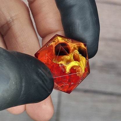 Red translucent resin D20 dice with a gold skull embedded inside, held between black-gloved fingers. Handmade gothic RPG dice for Dungeons & Dragons and dark fantasy tabletop gaming
