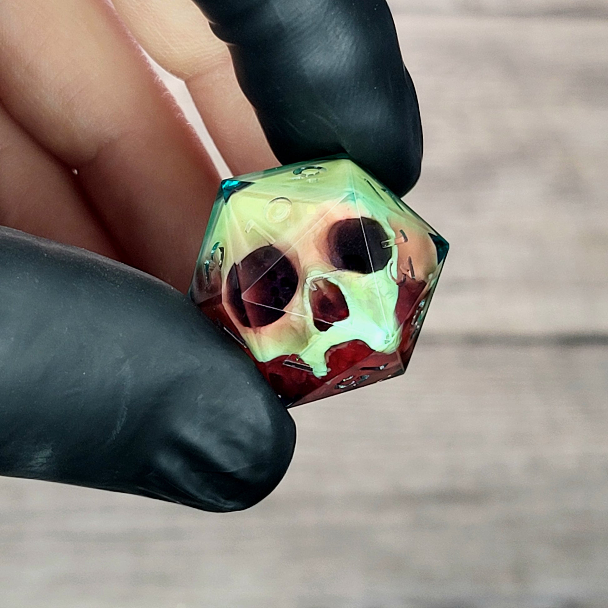 Handmade translucent resin D20 dice with embedded skull design in neon green and deep red colors, held between black-gloved fingers. Unique gothic RPG dice for Dungeons & Dragons and tabletop gaming