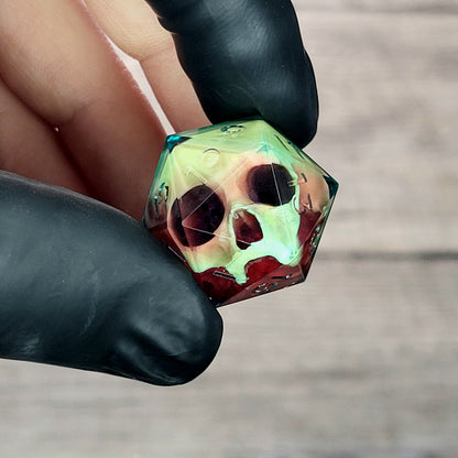 Handmade translucent resin D20 dice with embedded skull design in neon green and deep red colors, held between black-gloved fingers. Unique gothic RPG dice for Dungeons & Dragons and tabletop gaming