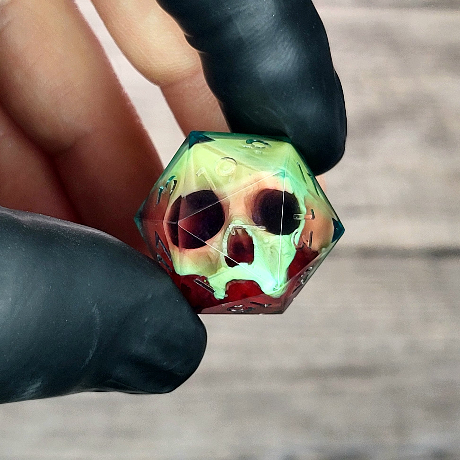 Handmade translucent resin D20 dice with embedded skull design in neon green and deep red colors, held between black-gloved fingers. Unique gothic RPG dice for Dungeons & Dragons and tabletop gaming