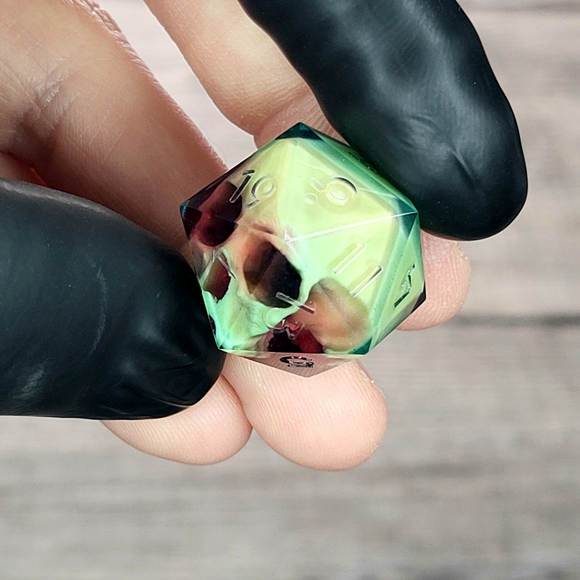 Handmade translucent resin D20 dice with embedded skull design in neon green and deep red colors, held between black-gloved fingers. Unique gothic RPG dice for Dungeons & Dragons and tabletop gaming