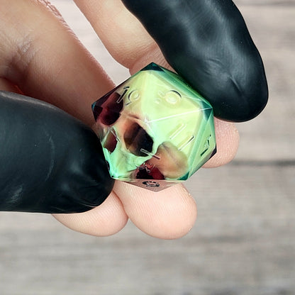 Handmade translucent resin D20 dice with embedded skull design in neon green and deep red colors, held between black-gloved fingers. Unique gothic RPG dice for Dungeons & Dragons and tabletop gaming