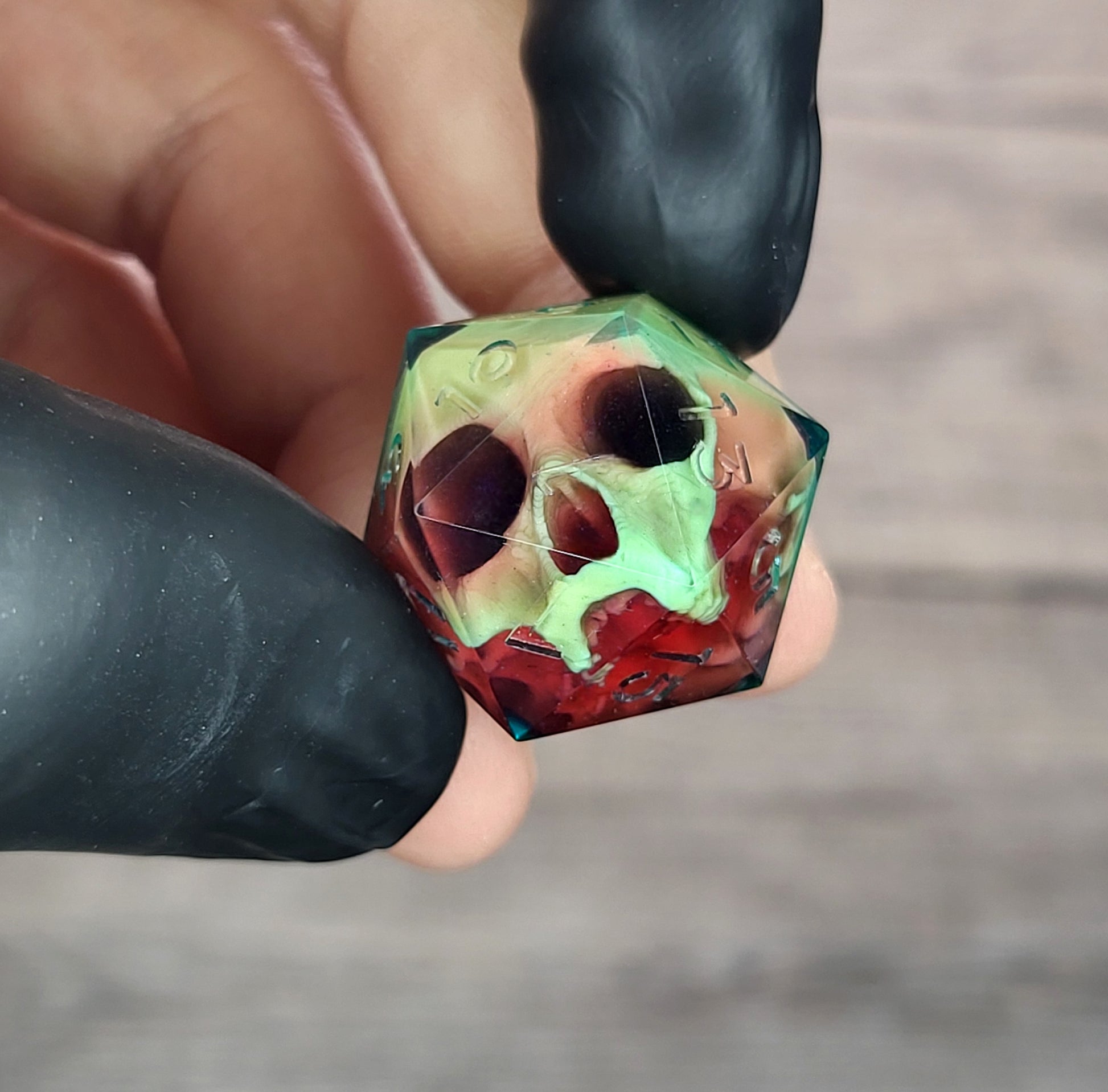 Handmade translucent resin D20 dice with embedded skull design in neon green and deep red colors, held between black-gloved fingers. Unique gothic RPG dice for Dungeons & Dragons and tabletop gaming