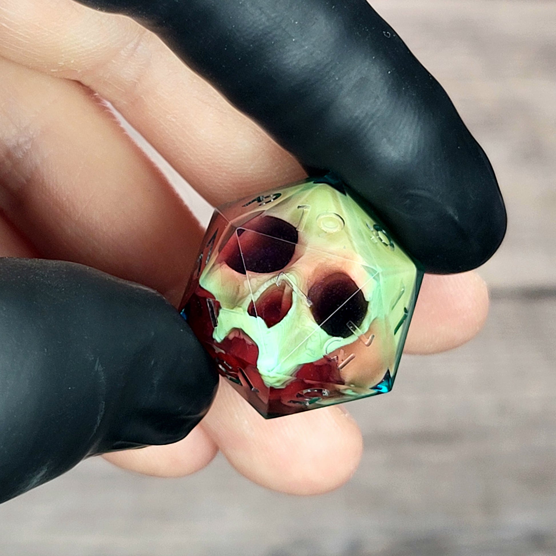 Handmade translucent resin D20 dice with embedded skull design in neon green and deep red colors, held between black-gloved fingers. Unique gothic RPG dice for Dungeons & Dragons and tabletop gaming
