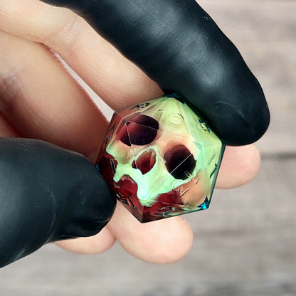 Handmade translucent resin D20 dice with embedded skull design in neon green and deep red colors, held between black-gloved fingers. Unique gothic RPG dice for Dungeons & Dragons and tabletop gaming