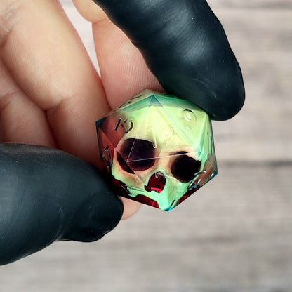 Handmade translucent resin D20 dice with embedded skull design in neon green and deep red colors, held between black-gloved fingers. Unique gothic RPG dice for Dungeons & Dragons and tabletop gaming